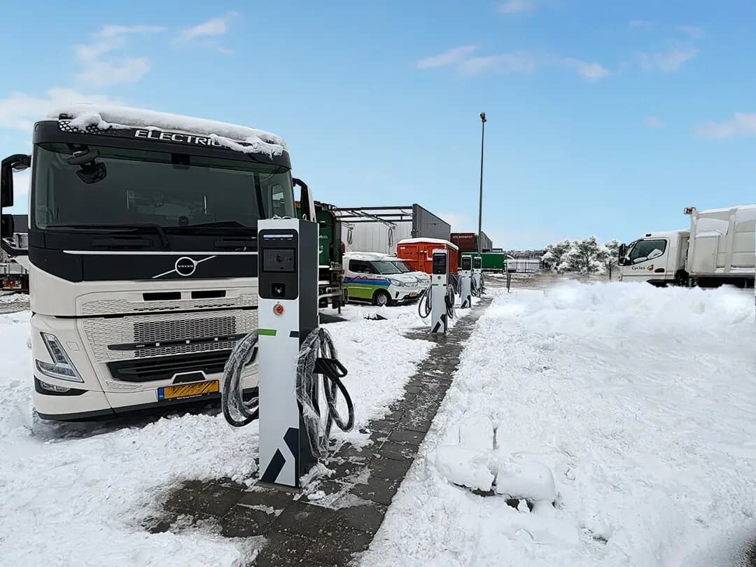 Witte elektrische Volvo vrachtwagen geparkeerd bij een laadpaal in een dik pak witte sneeuw.